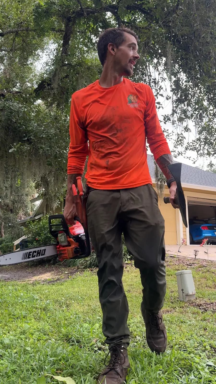Alex in a Squirrel’s Friend shirt carrying a chainsaw across a shaded yard