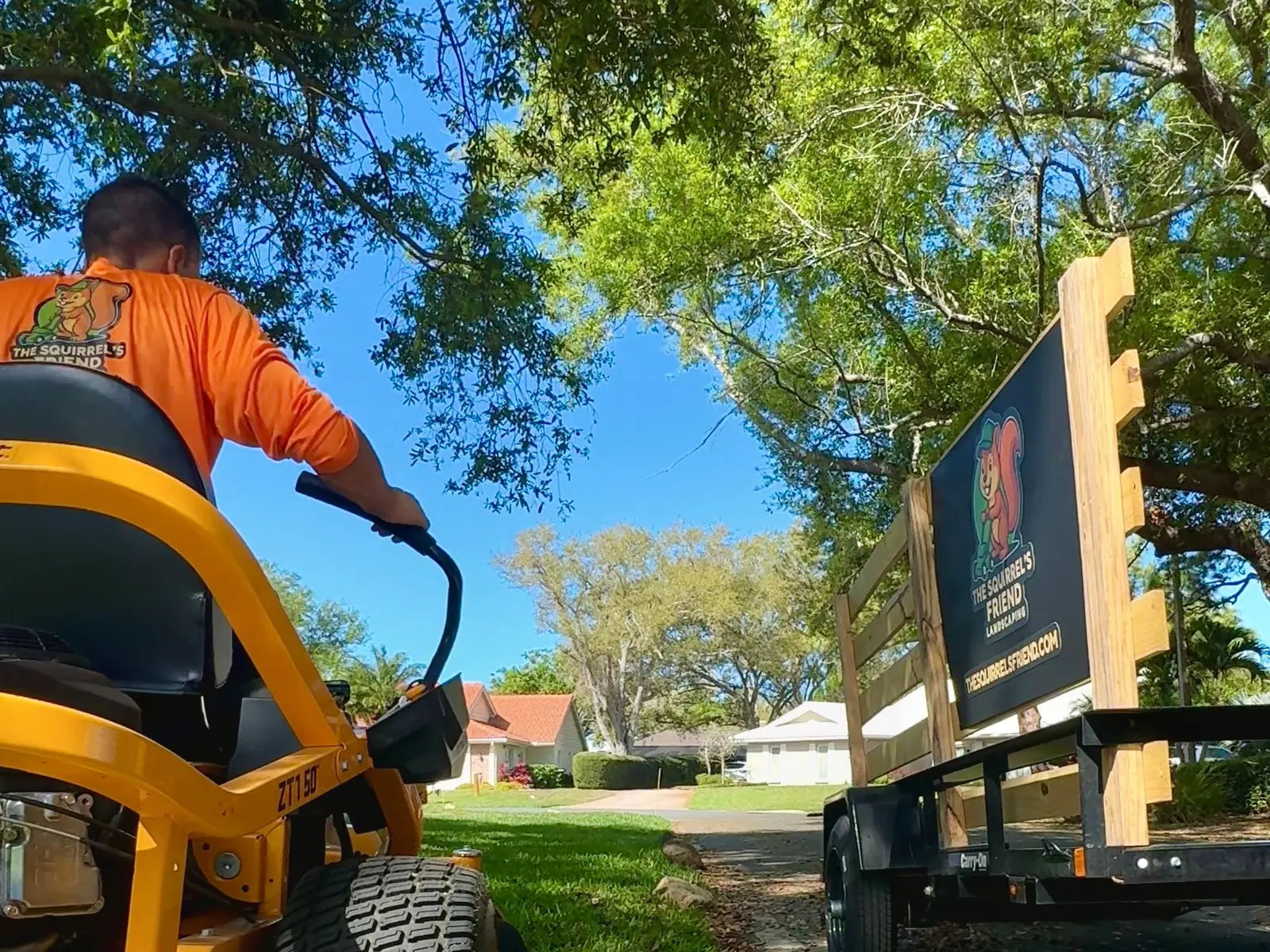 Alex mowing in a Squirrel’s Friend shirt, with the branded trailer sign beside him